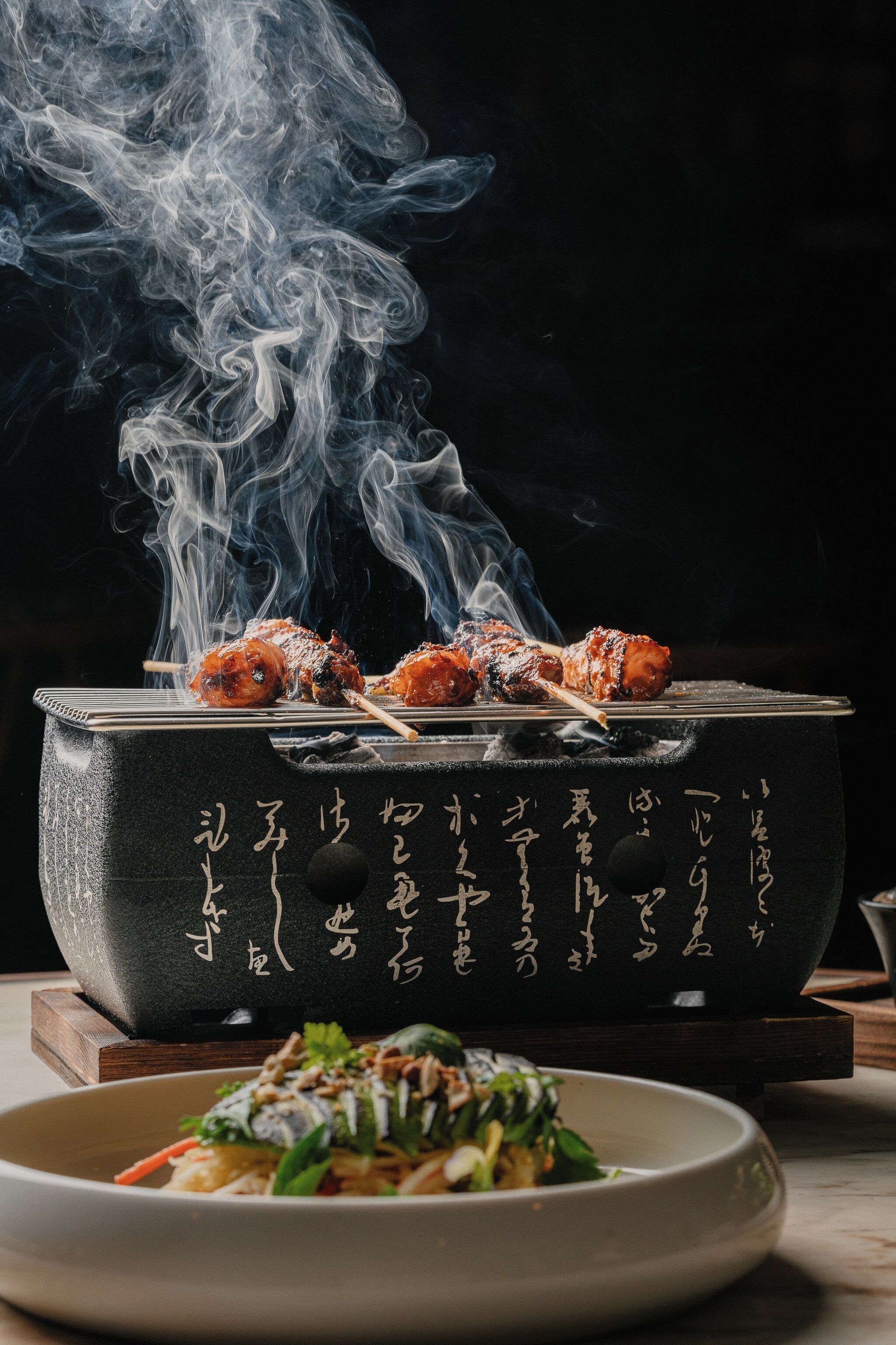 Una parrilla japonesa tradicional con brochetas de comida, un tazón de ensalada y humo elevándose de la parrilla.