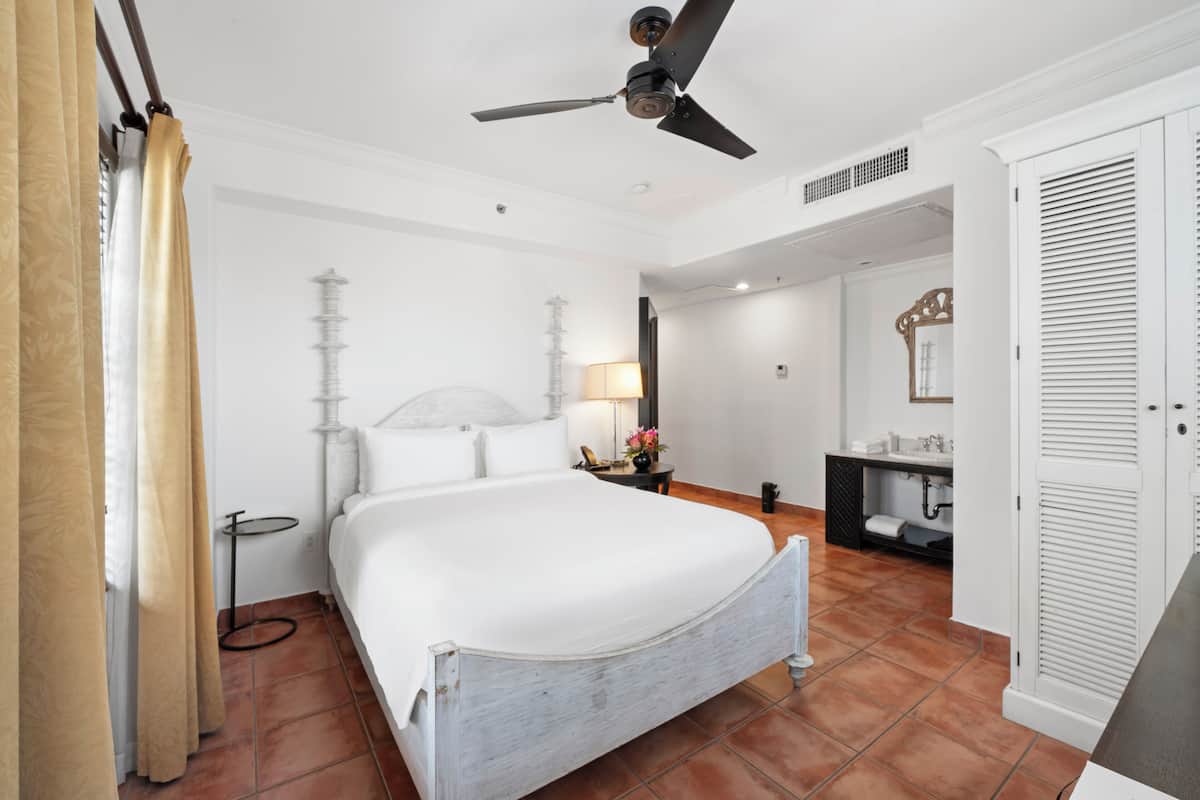 A hotel room with a bed, a ceiling fan, a bedside table with a lamp, a bathroom with a sink and mirror, and a wardrobe.