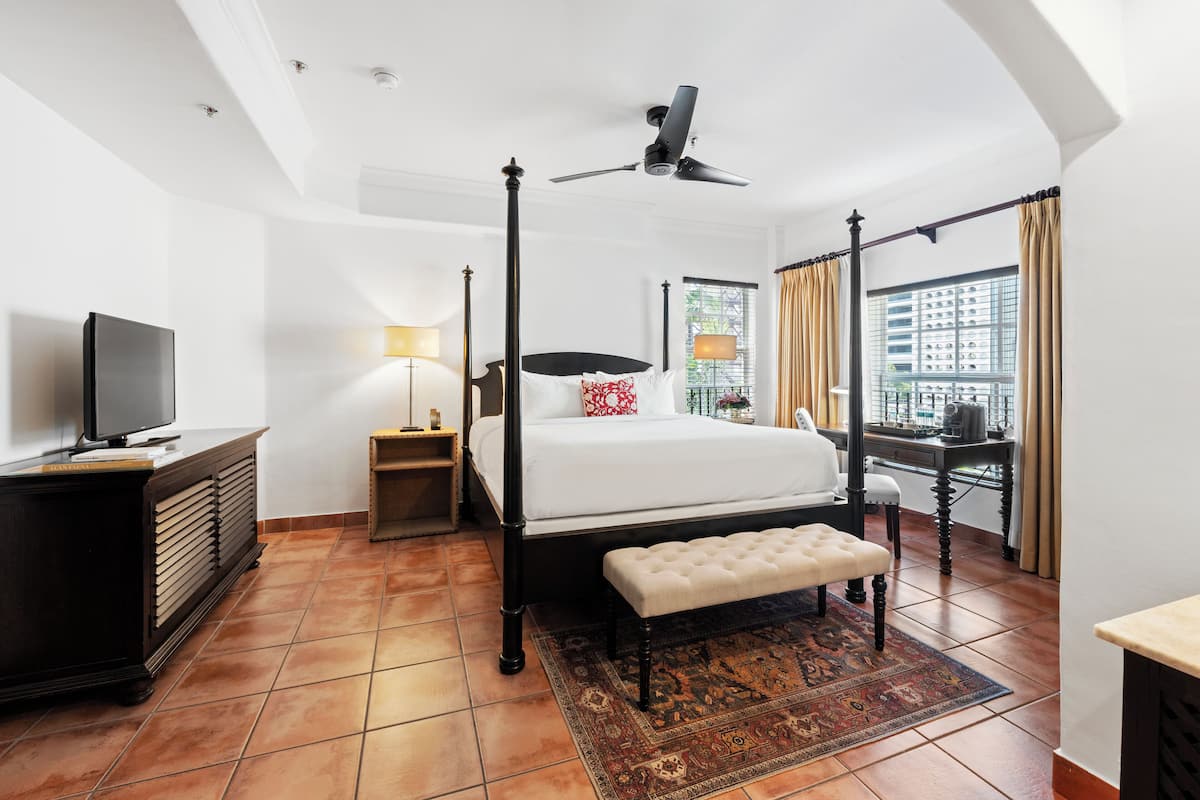 A hotel room with a four-poster bed, a television, a desk, and a view of the city through large windows.