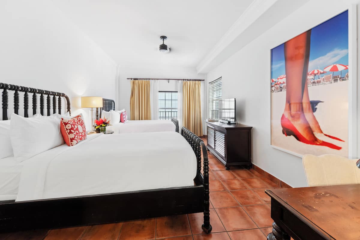 A hotel room with a large bed, a desk, and a chair. There is a window with curtains, a painting on the wall, and a ceiling fan.