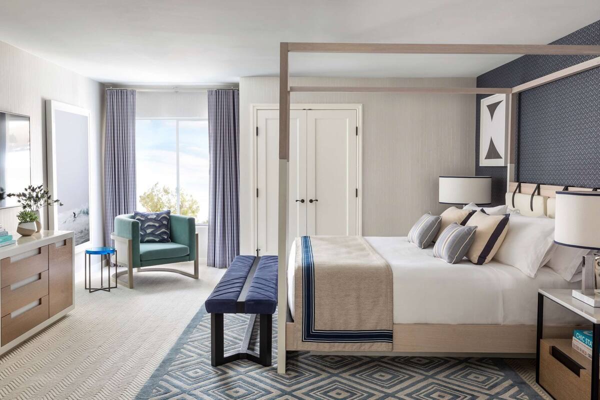 Una habitación de hotel con cama, mesitas de noche, escritorio, silla, televisor y ventana con cortinas.