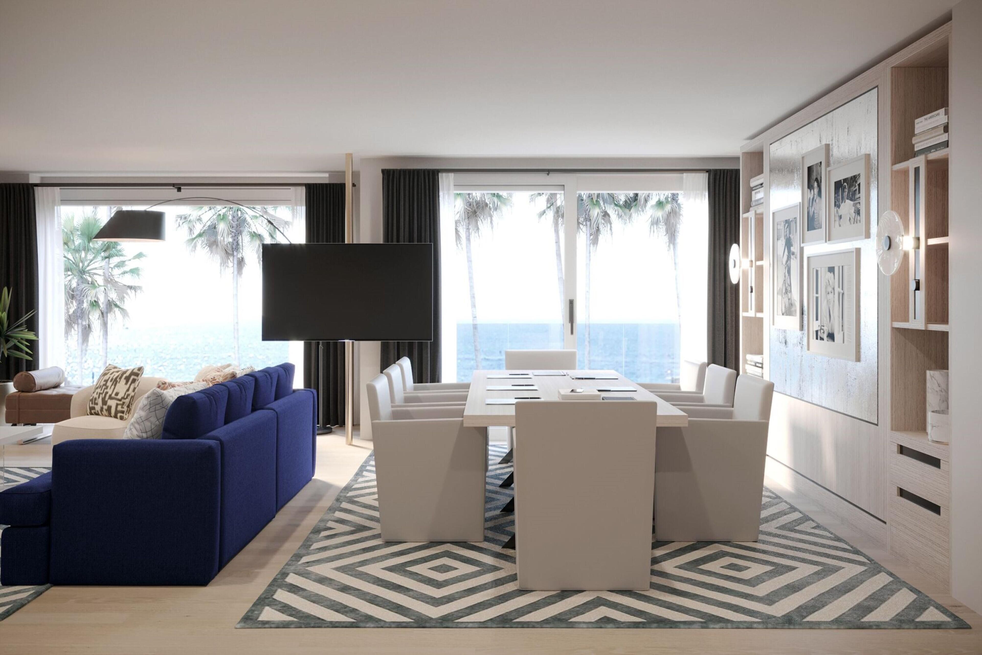 A modern living room with a blue sofa, a dining table with chairs, a flat-screen TV, and a view of the ocean through large windows.