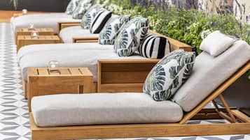 A row of wooden lounge chairs with cushions, arranged on a patterned floor, with a garden and lavender plants in the background.