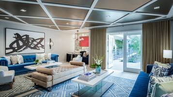 A modern living room with a blue sofa, a beige sofa, and a coffee table with books. There is a large abstract painting on the wall and a view of the outdoors through a sliding door.