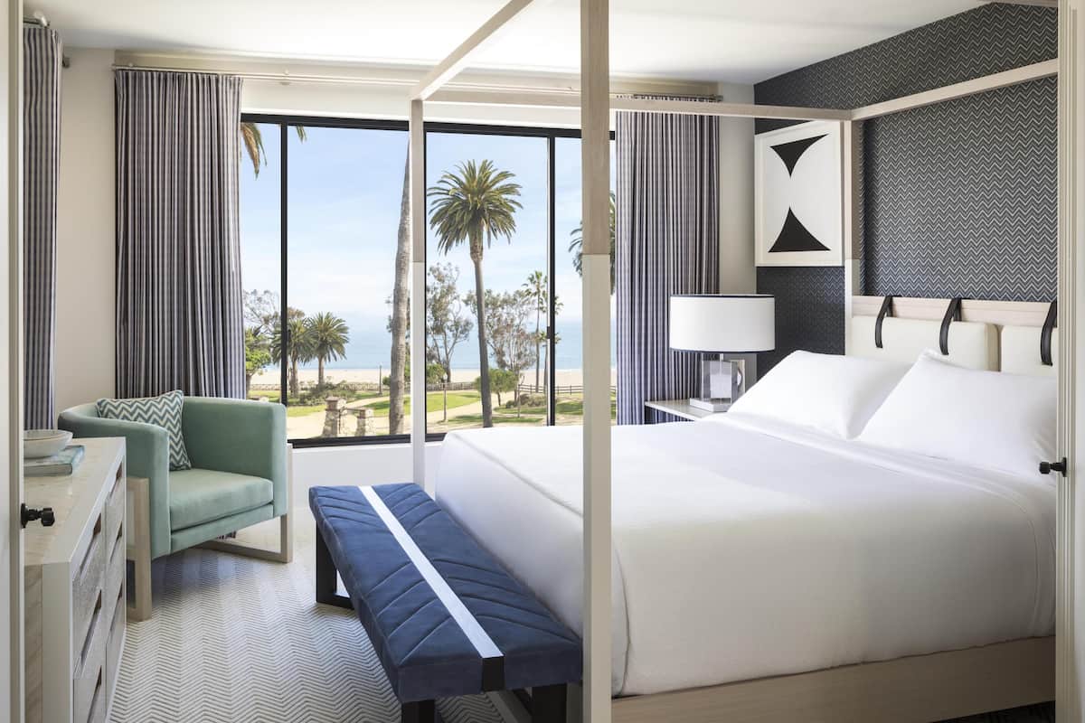 Una cama con dosel con vista al mar, un banco, un escritorio con una silla y una mesita de noche con lámpara.