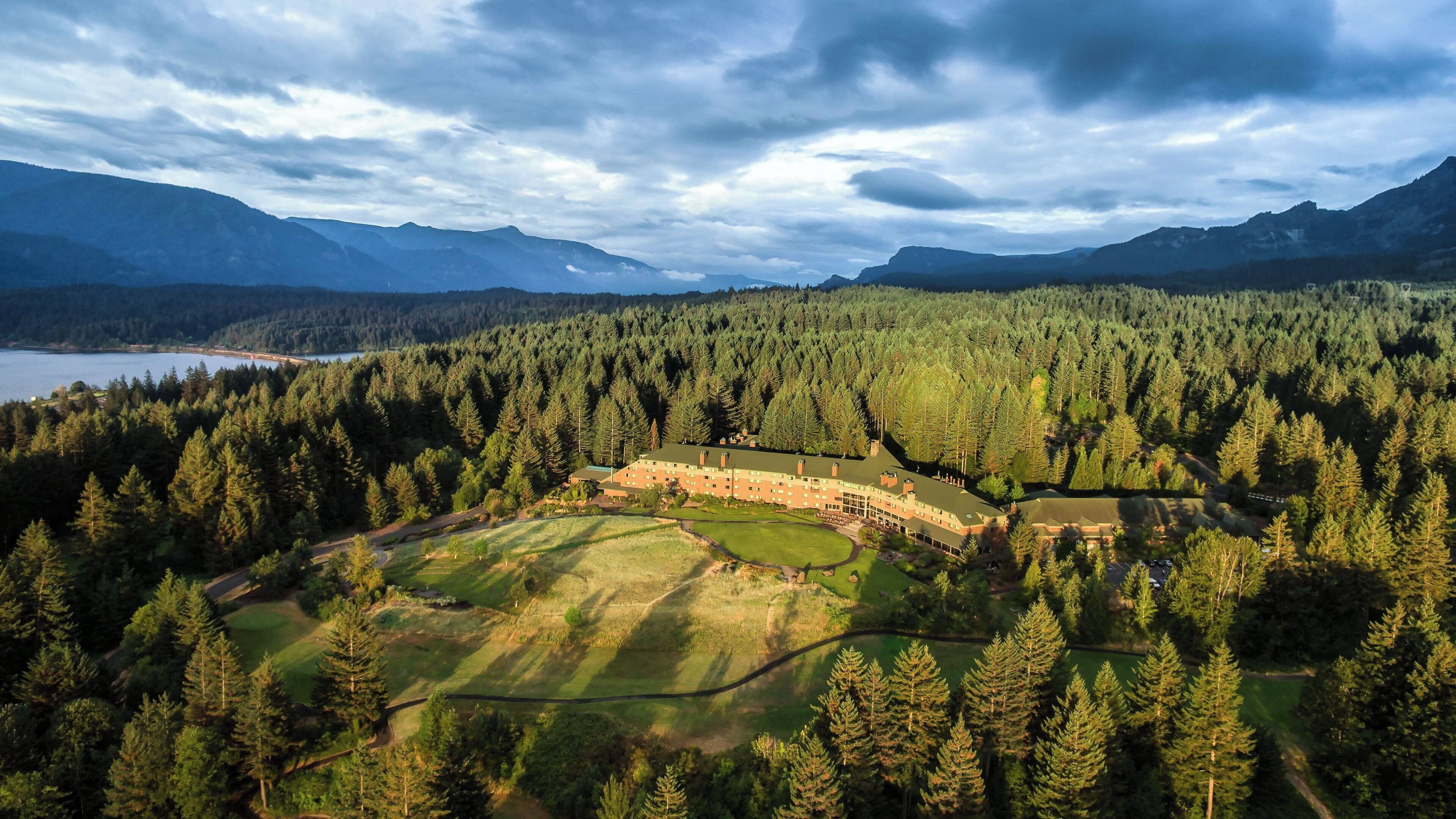 Un hotel rodeado de un bosque denso, con un campo de golf y montañas al fondo.