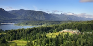 Un chalet avec un grand bâtiment, entouré d’une forêt dense et de montagnes. Un pont enjambe un lac au loin.
