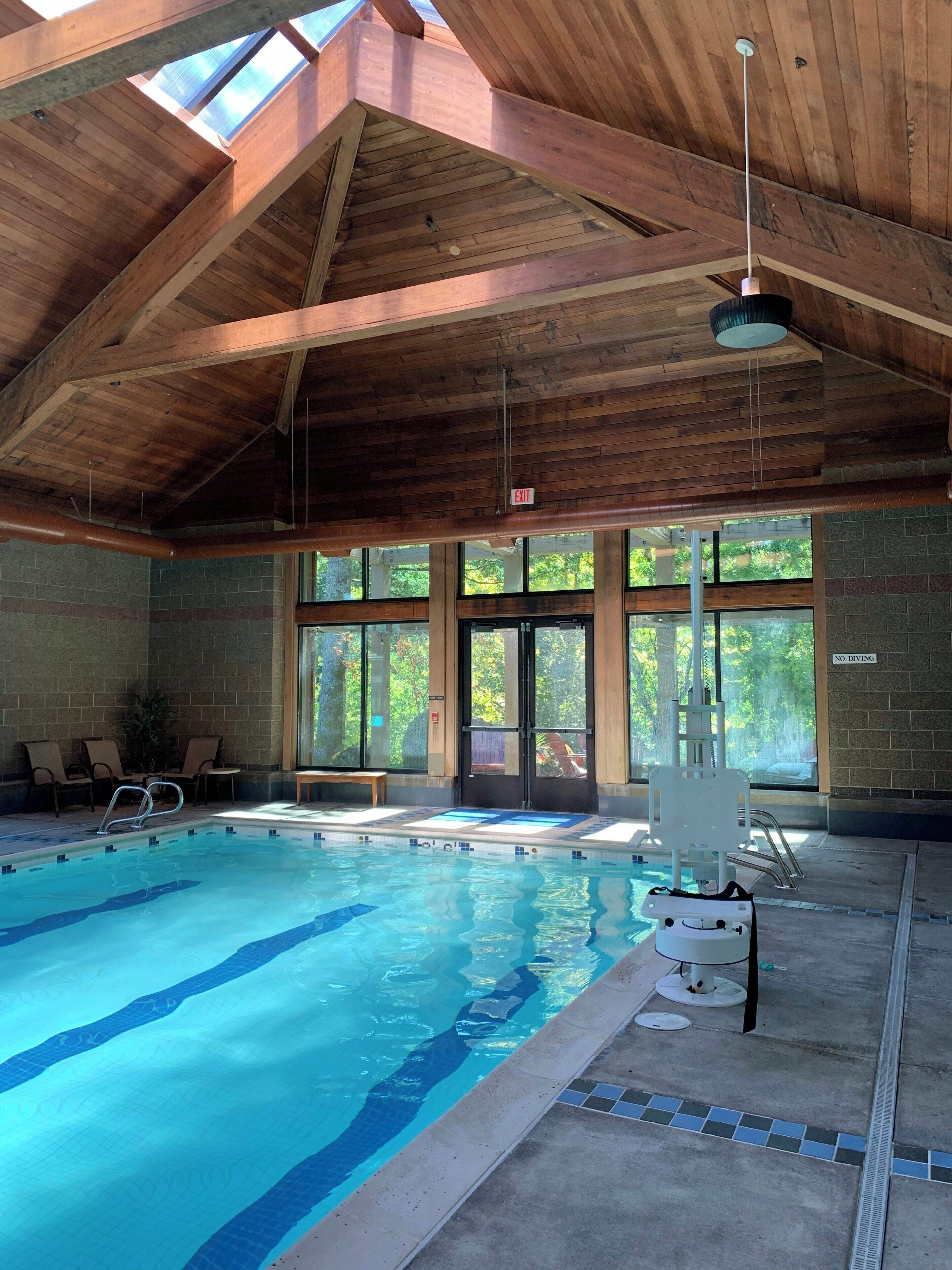 An indoor swimming pool with clear blue water, wooden beams on the ceiling, and a bench area with chairs.