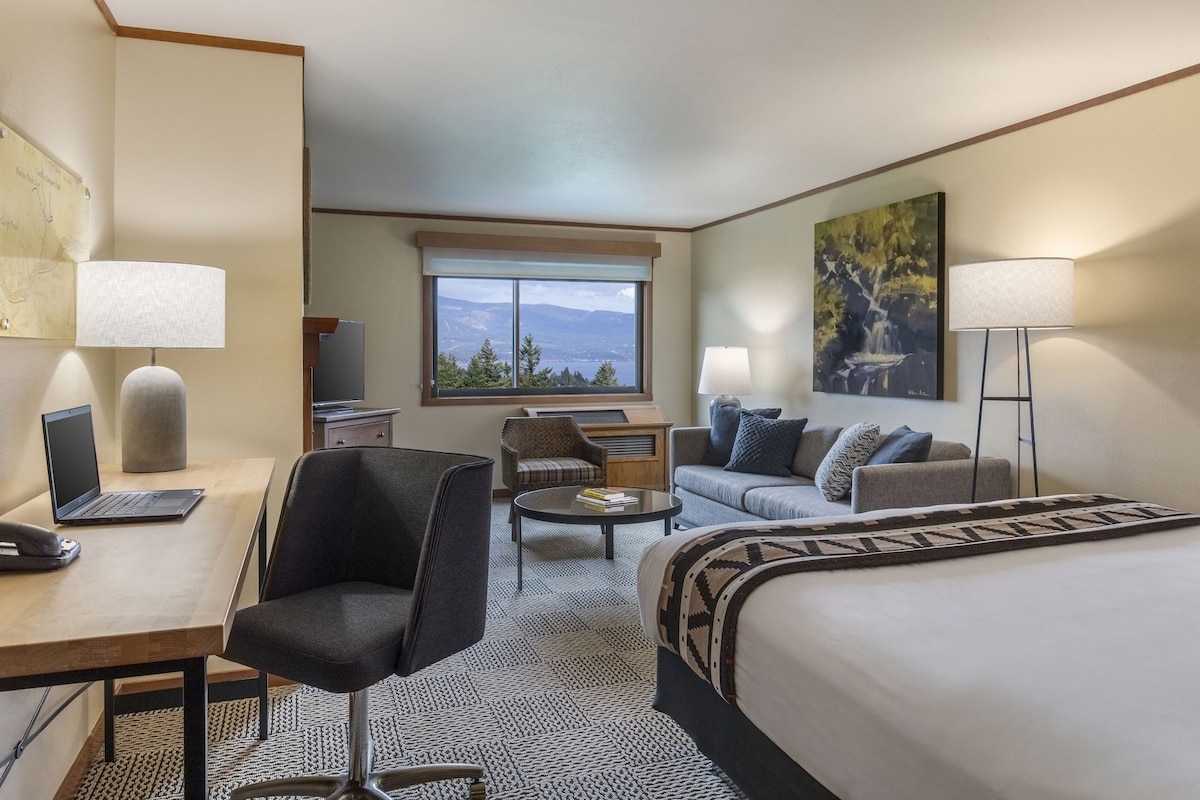 A hotel room with a bed, a desk with a laptop, a chair, a sofa, a coffee table, and a window with a view.