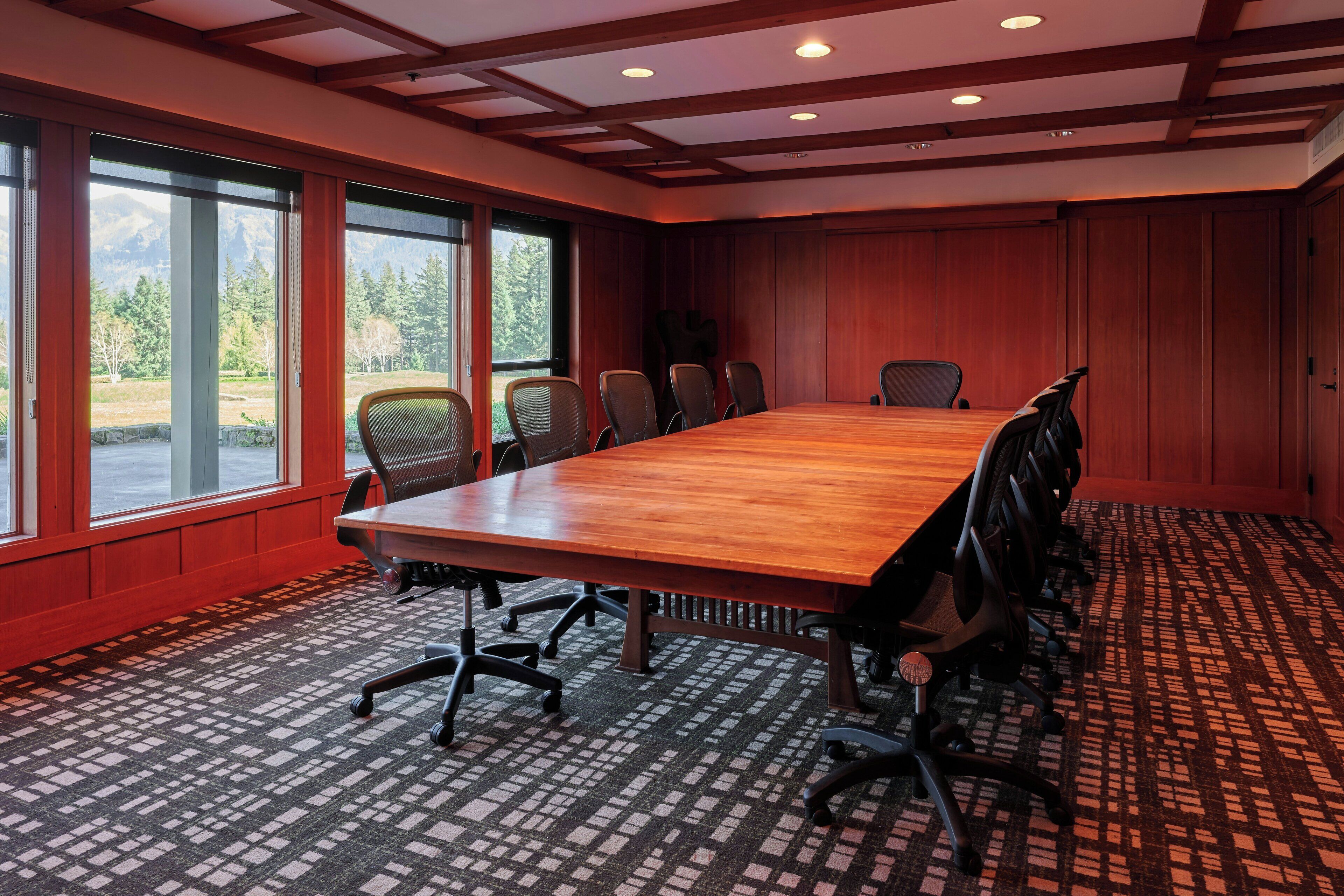 A conference room with a large wooden table, multiple chairs, and large windows offering a view of trees and mountains.