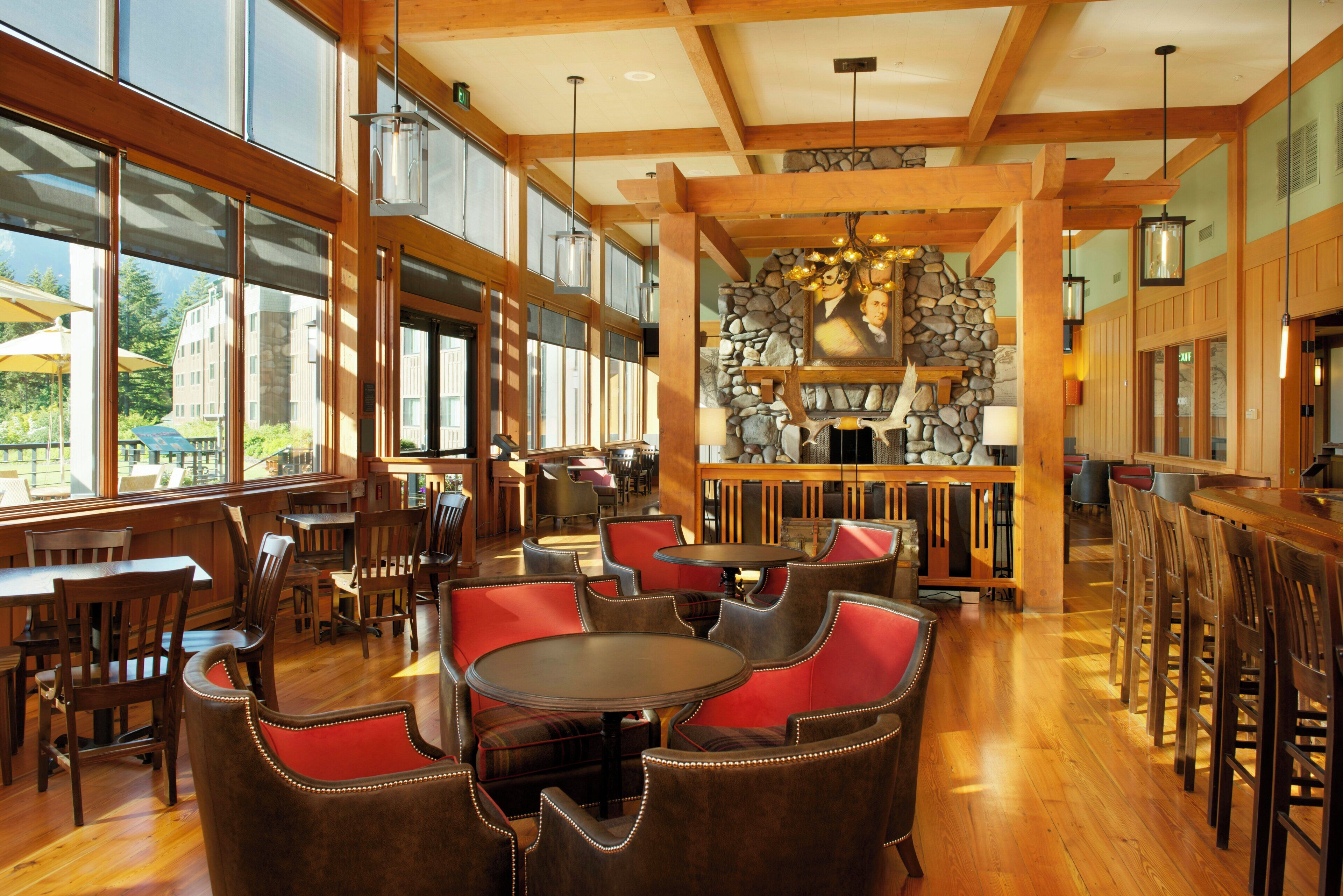 A spacious indoor area with wooden flooring, multiple tables and chairs, and large windows allowing natural light.
