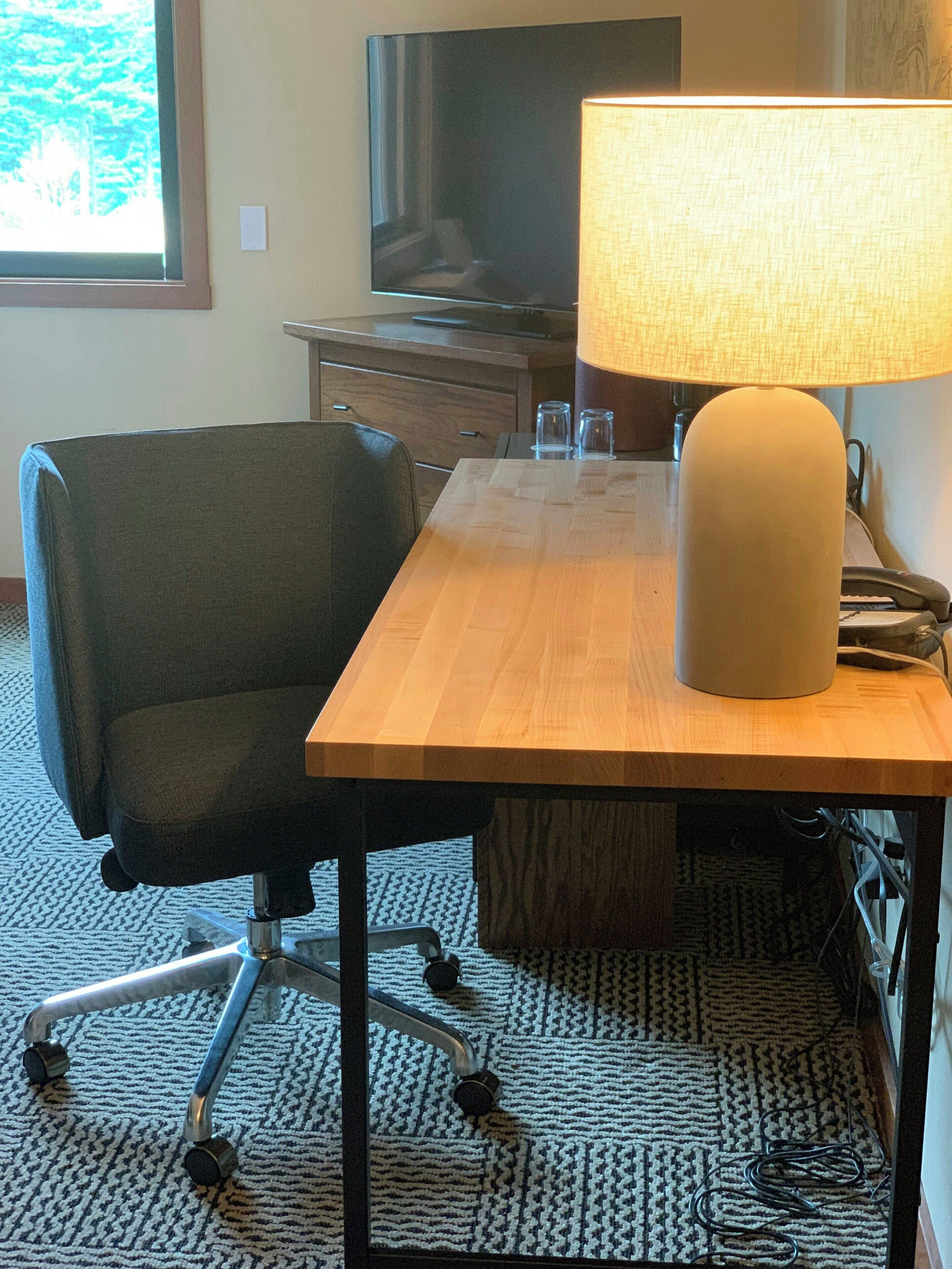 A hotel room with a wooden desk, a chair, a lamp, a television, and a window with a view of trees.