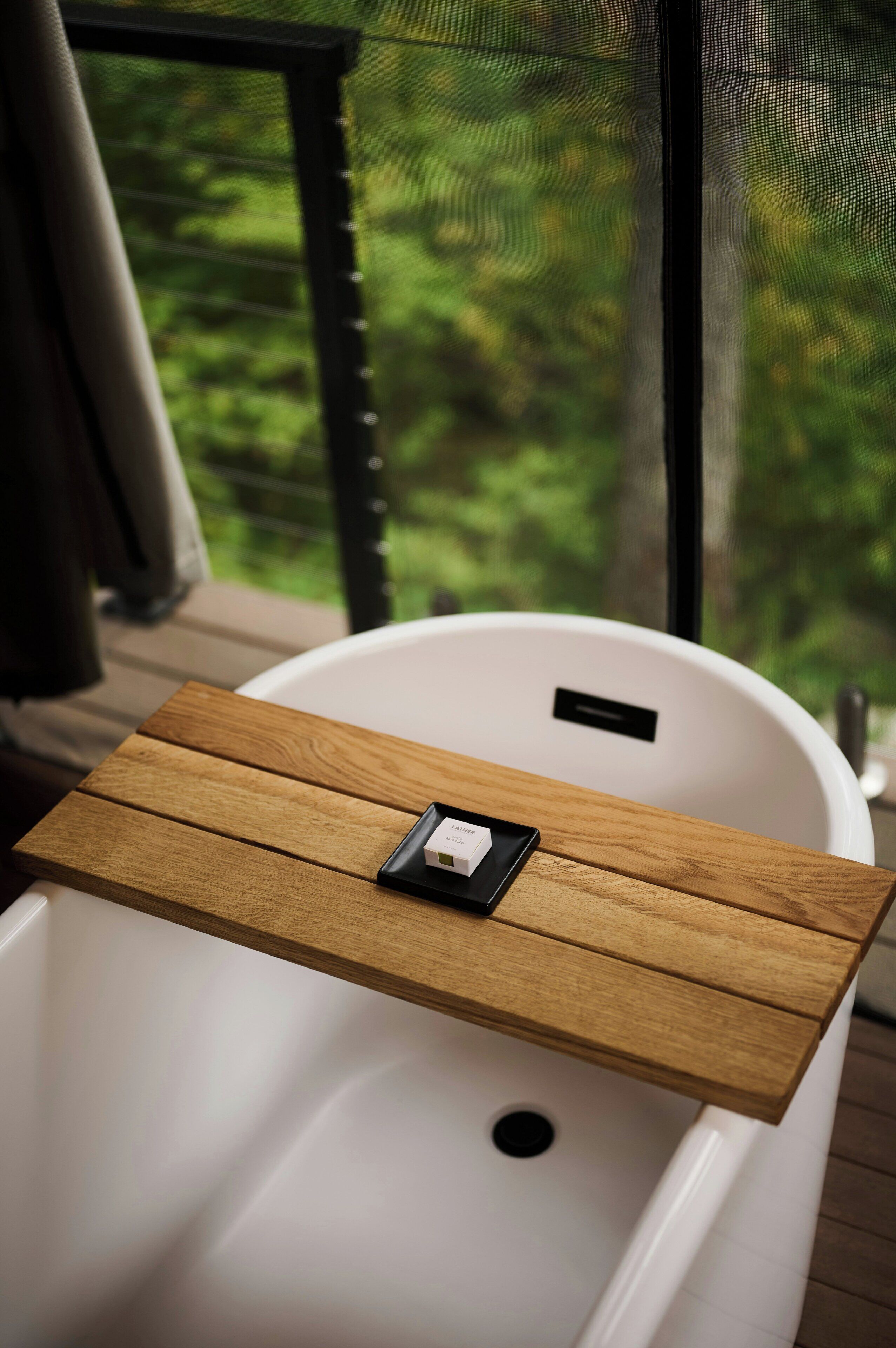 A white bathtub with a wooden tray on top, a small box labeled 'LATHER,' and a view of greenery through a window.
