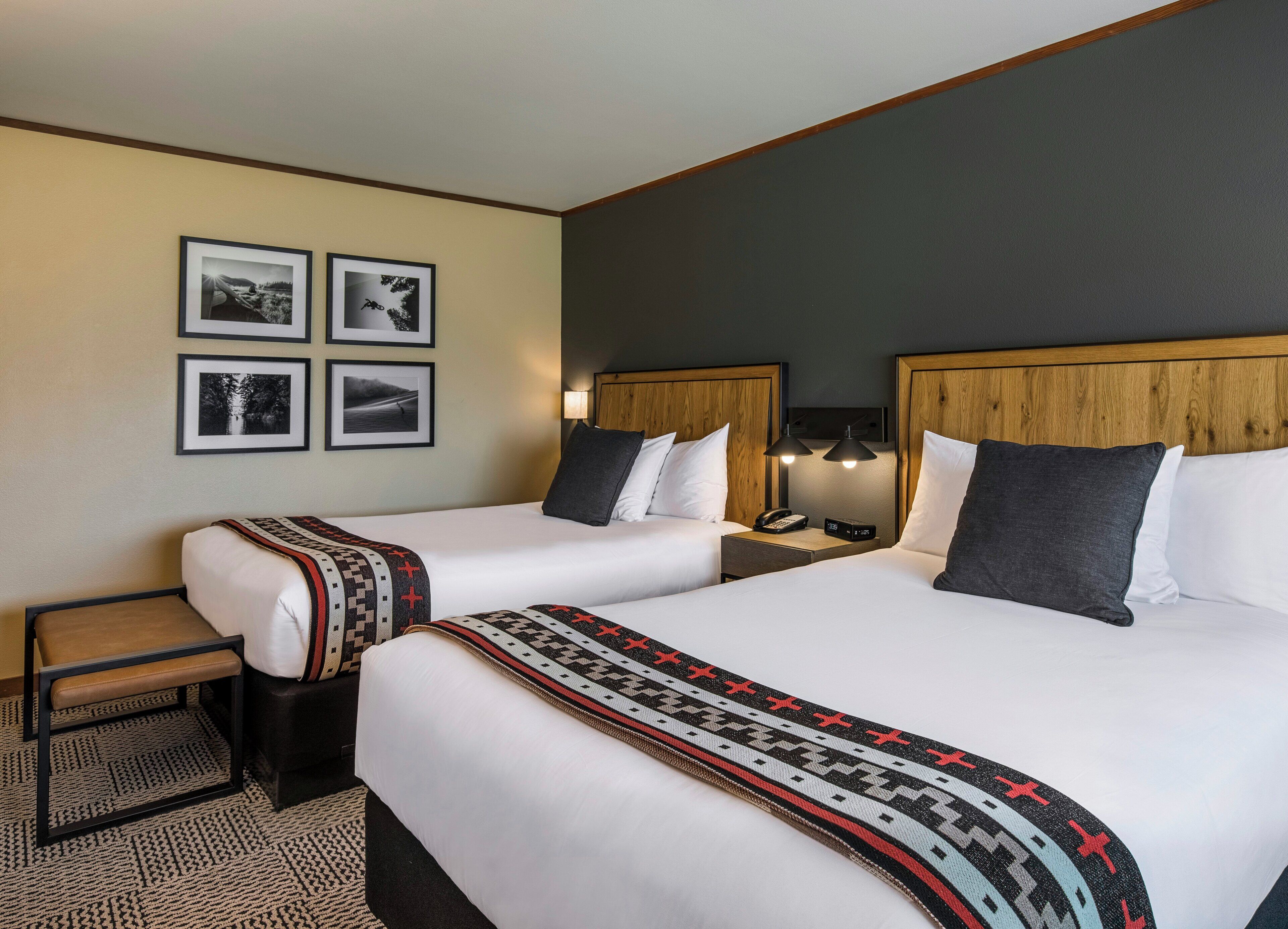 A hotel room with two beds, a nightstand with a lamp, a small table, and a bench. There are four framed pictures on the wall.