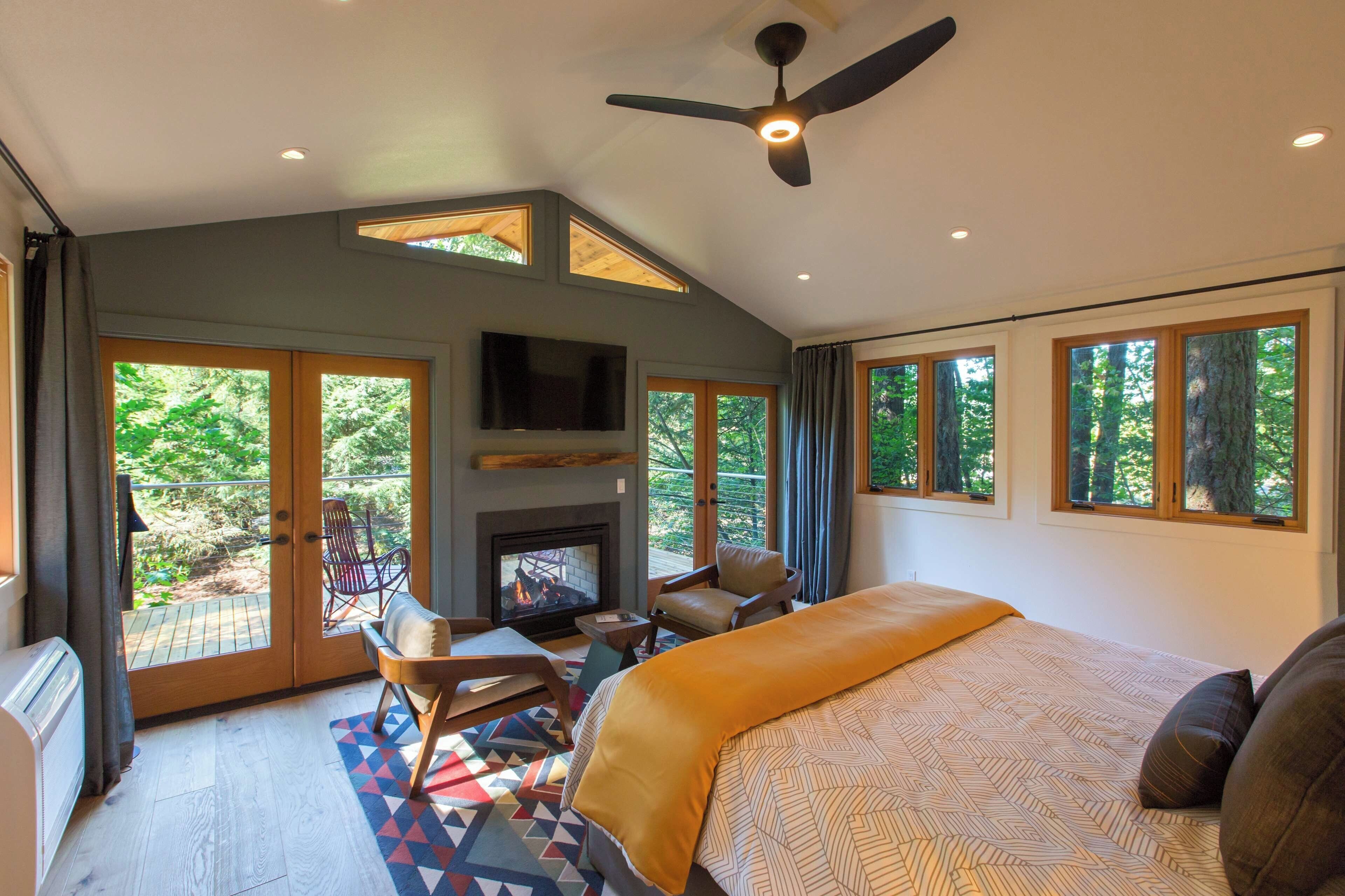 A bedroom with a large bed, a fireplace, a ceiling fan, and large windows.
