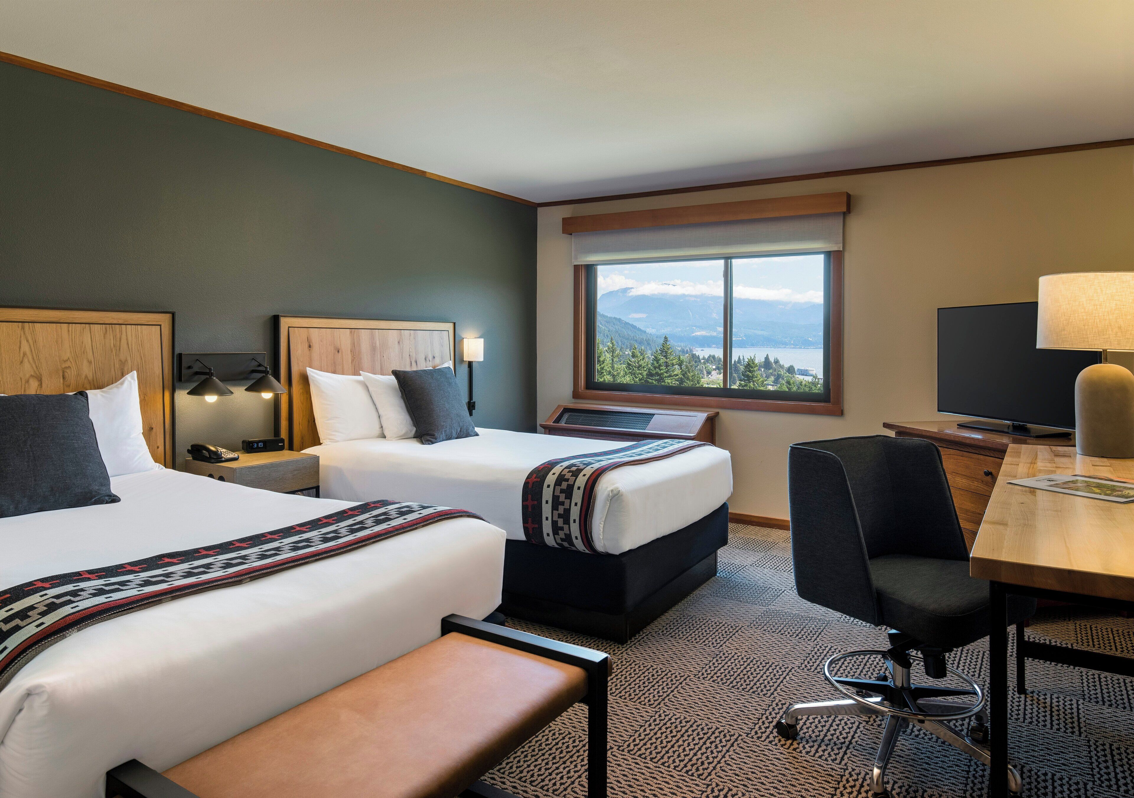 A hotel room with two beds, a desk, a chair, and a large window with a mountain view.