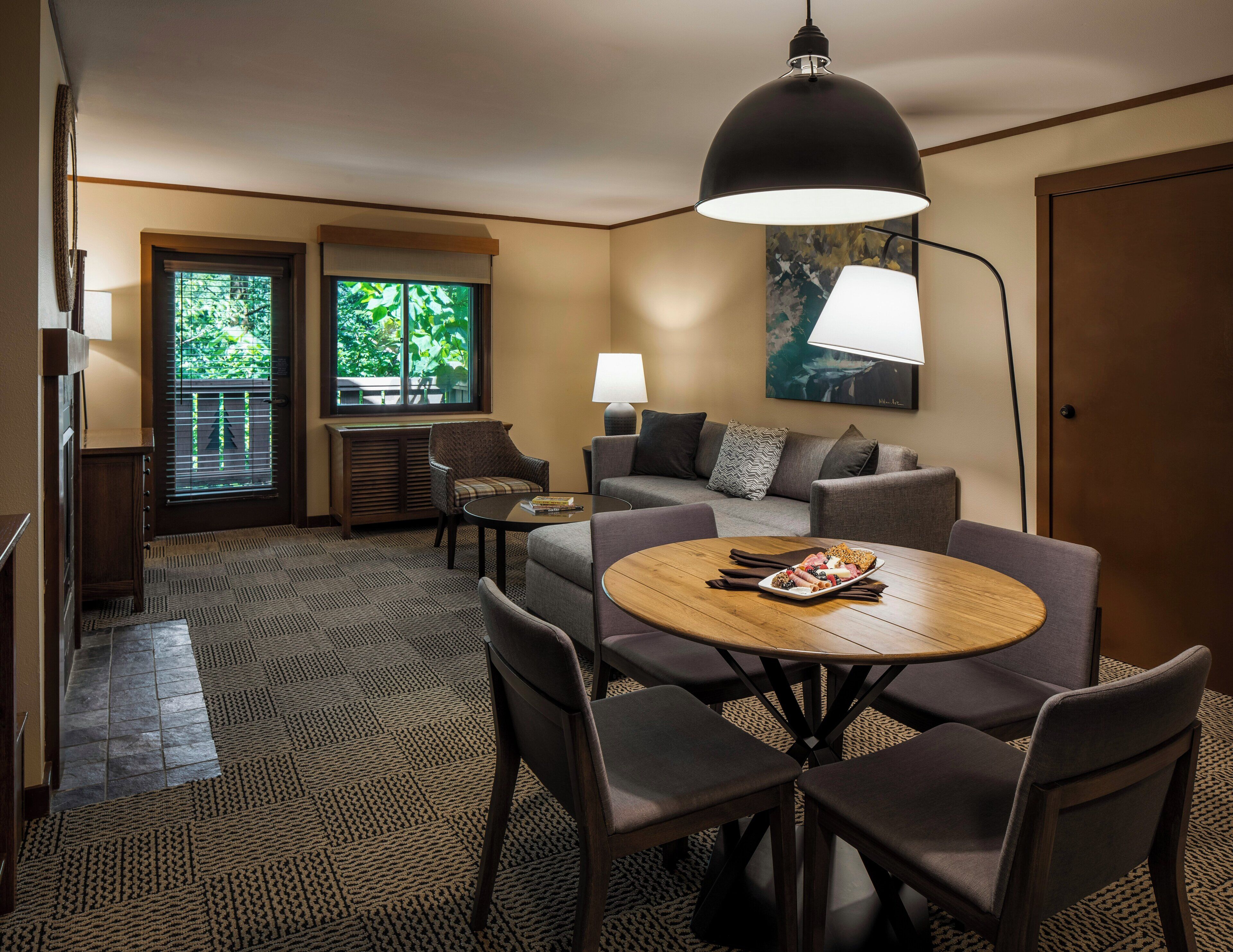 Habitación de hotel con mesa de comedor, sillas, sofá, lámpara, un cuadro y ventana con persianas.
