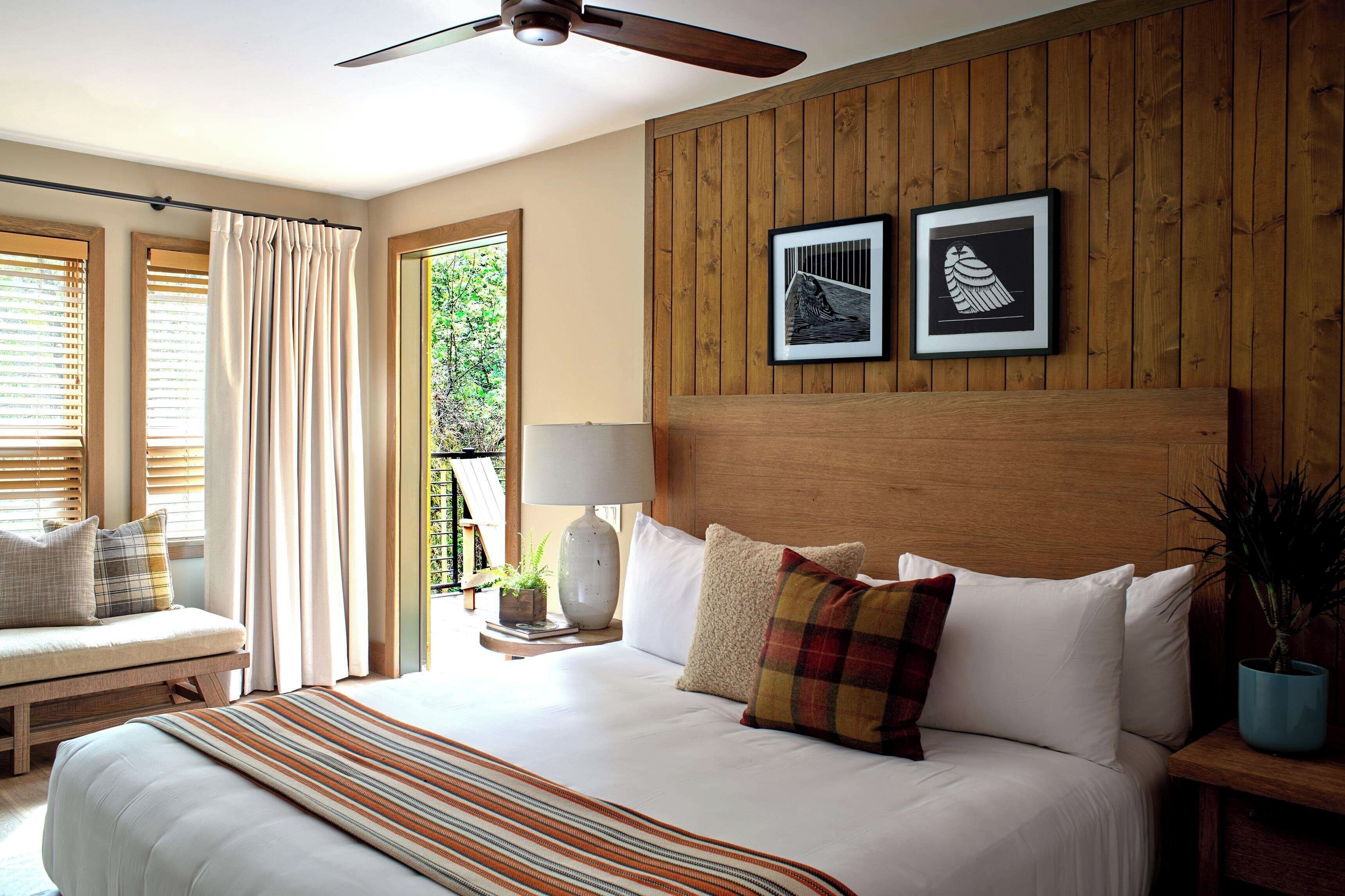 A bedroom with a large bed, a ceiling fan, wooden panel walls, and two framed artworks.