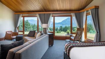 A hotel room with a large bed, a sofa, a chair, and a small table. There are multiple windows offering a view of the outdoors.