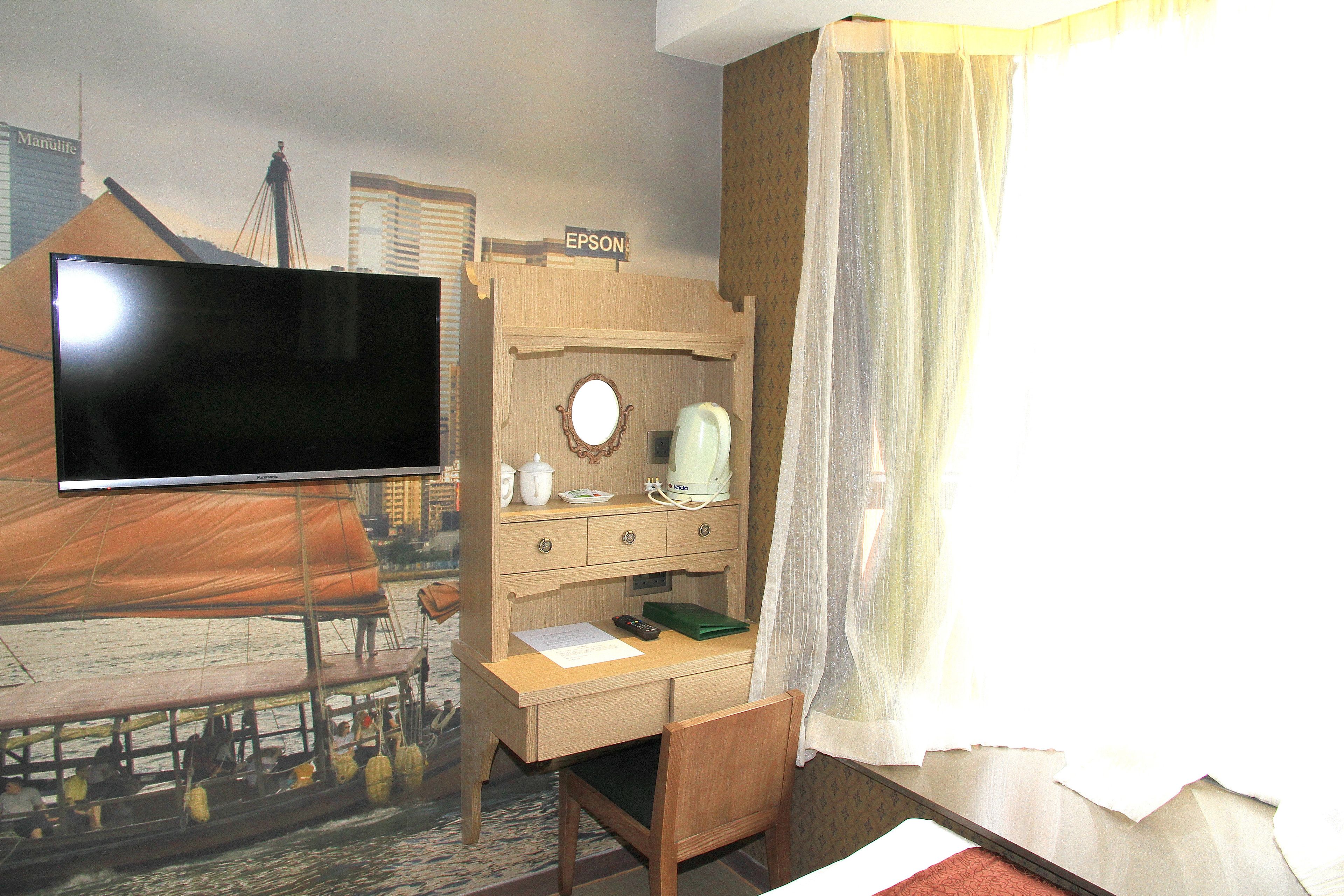 Een hotelkamer met een grote flatscreen-tv, een houten bureau, een stoel, een klein tafeltje, een bed met een witte sprei en een raam met dunne gordijnen.