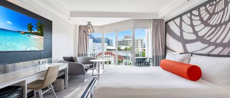 A hotel room with a large bed, a desk with a chair, a TV, and a balcony with a view of the city and palm trees.