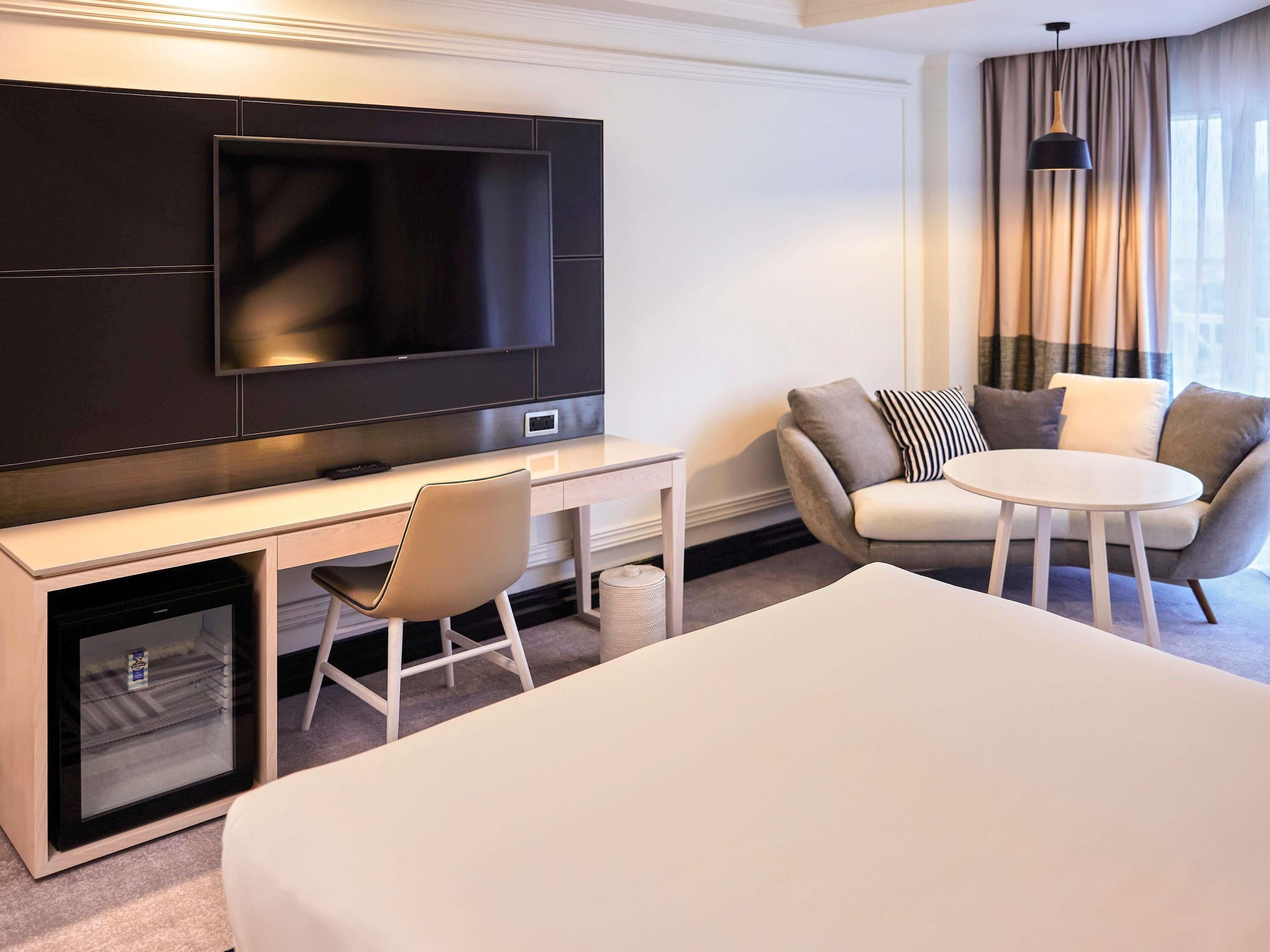 A hotel room with a large flat-screen TV mounted on the wall, a desk with a chair, a round table with two chairs, and a bed.