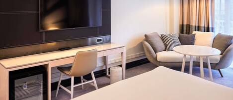A hotel room with a large flat-screen TV mounted on the wall, a desk with a chair, a round table with two chairs, and a bed.