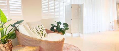 Una sala de estar moderna con un sofá curvo, un tapete redondo y amplias ventanas con persianas blancas.