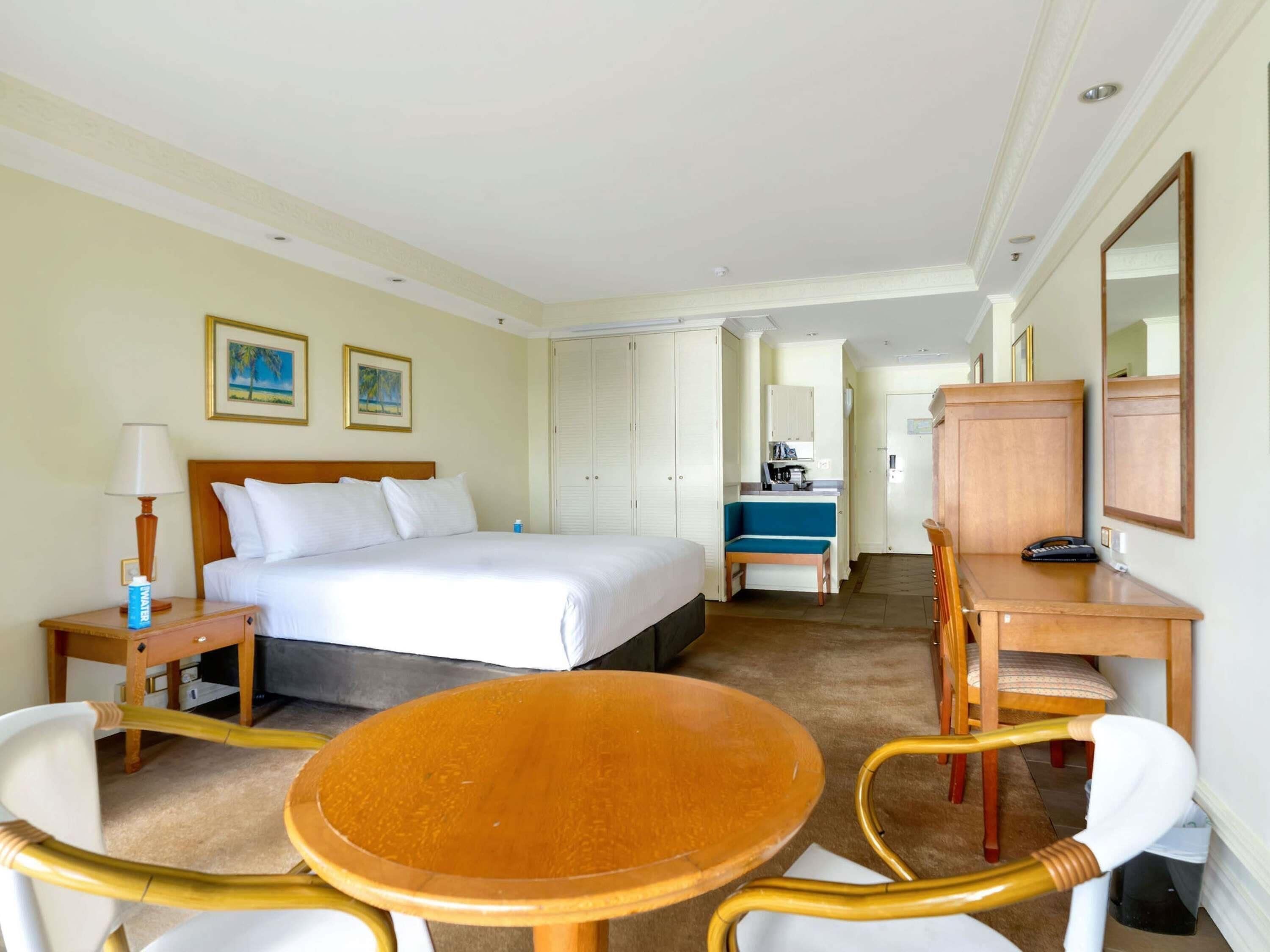 A hotel room with a bed, a desk with a chair, a small table, a mirror, and a kitchenette in the background.