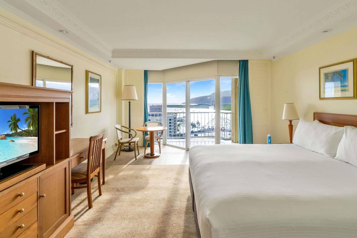 Une chambre d’hôtel comprenant un lit, un bureau avec une télévision, une chaise, une petite table, un balcon avec vue et une fenêtre avec des rideaux.