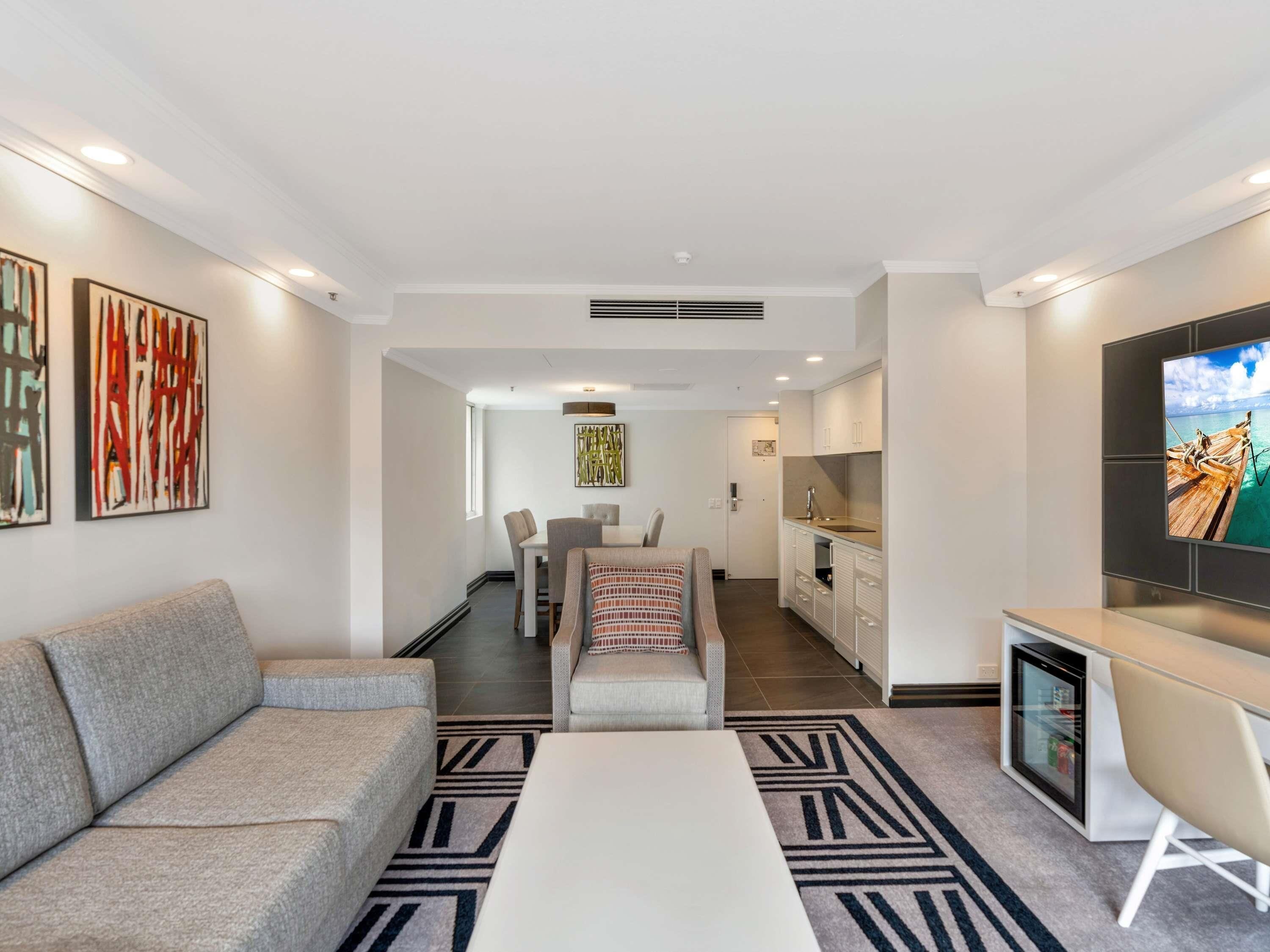 Una sala moderna con sofá, mesa de centro y televisor. Cuenta con una zona de cocina con armarios y refrigerador. El ambiente presenta una paleta de colores neutros.