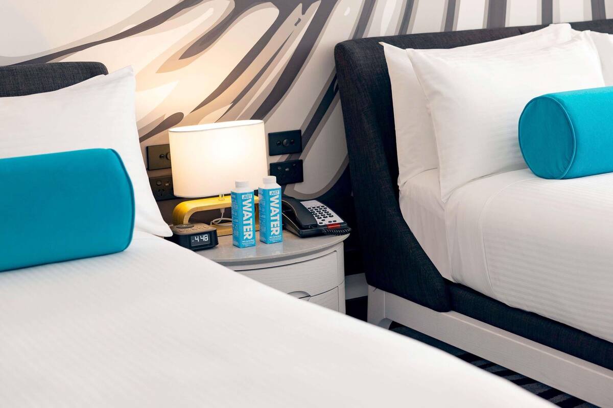 Une chambre d’hôtel avec deux lits, une table de chevet avec une lampe, un téléphone et deux bouteilles d’eau.
