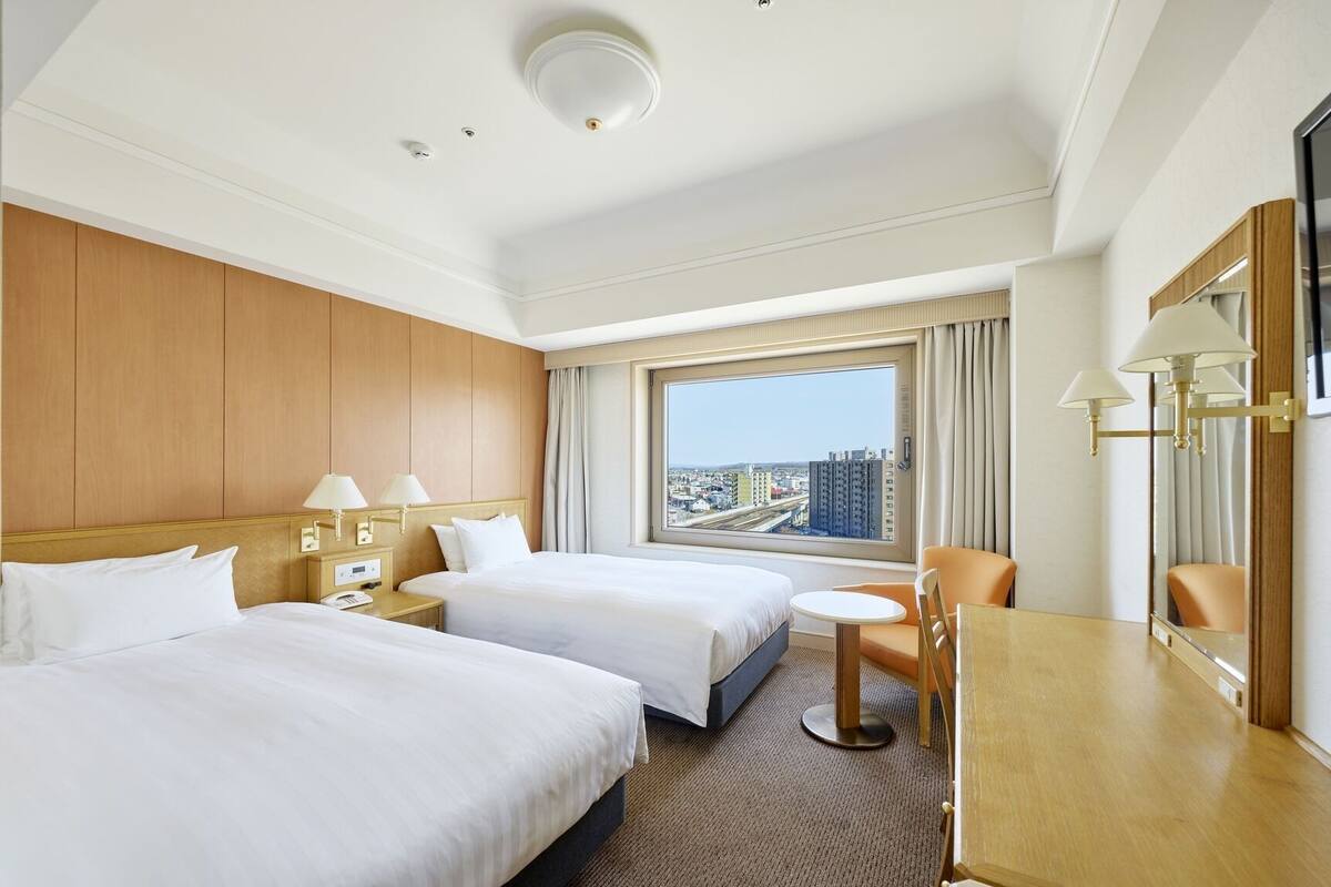 Une chambre d’hôtel avec deux lits, un bureau, une chaise, une grande fenêtre donnant sur la ville et un miroir.