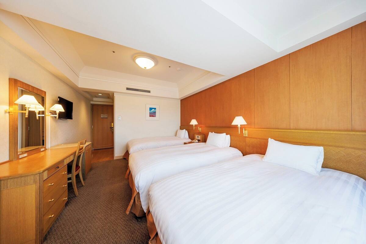 Une chambre d’hôtel avec trois lits simples, un bureau en bois avec une chaise, un grand miroir et deux lampes fixées au mur.