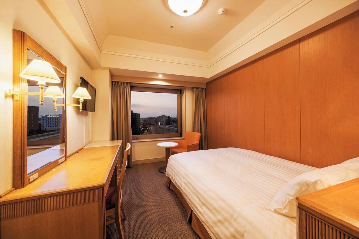 Une chambre d’hôtel équipée d’un lit, d’un bureau, d’une chaise, d’une petite table, d’un miroir et d’une fenêtre donnant sur la ville.
