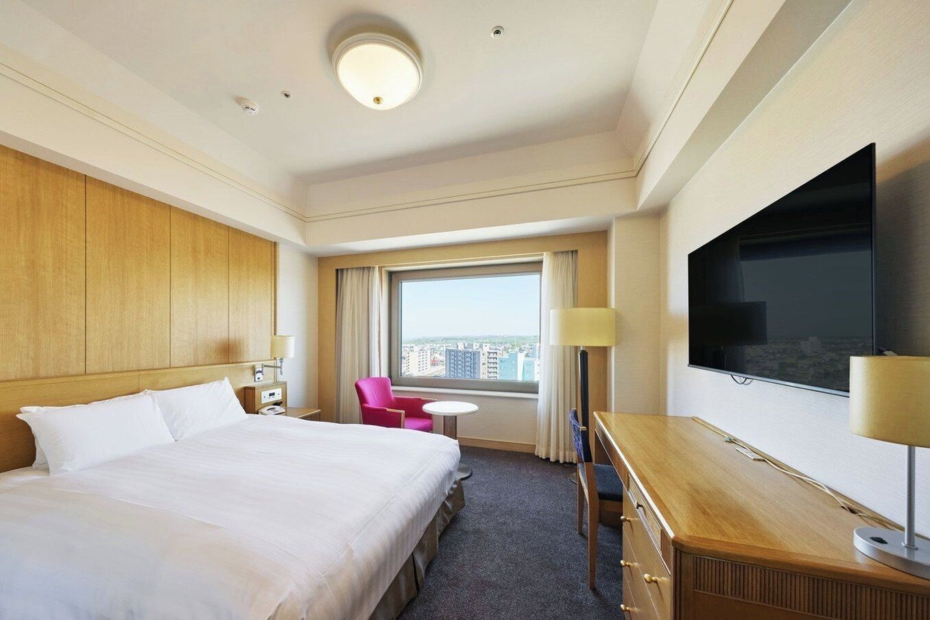 A hotel room with a large bed, a desk with a TV, a chair, a small table, and a window with a city view.