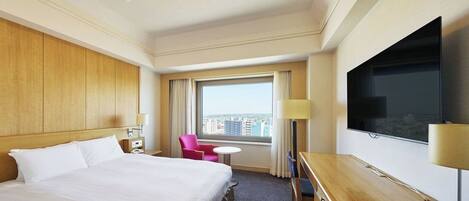 A hotel room with a large bed, a desk with a TV, a chair, a small table, and a window with a city view.