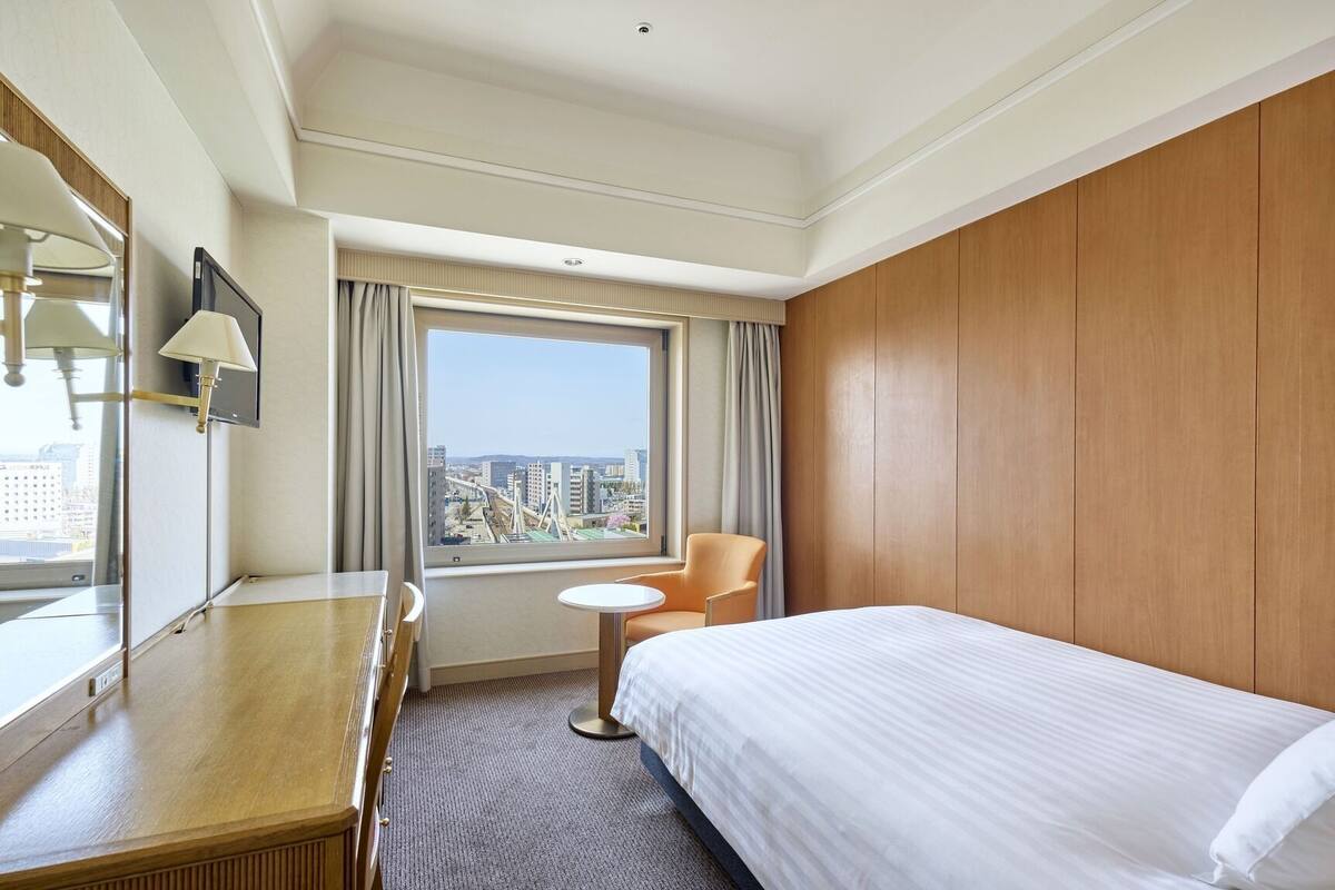 Une chambre d’hôtel comprenant un lit, un bureau, une chaise, une petite table, une fenêtre donnant sur la ville et des lambris en bois sur les murs.