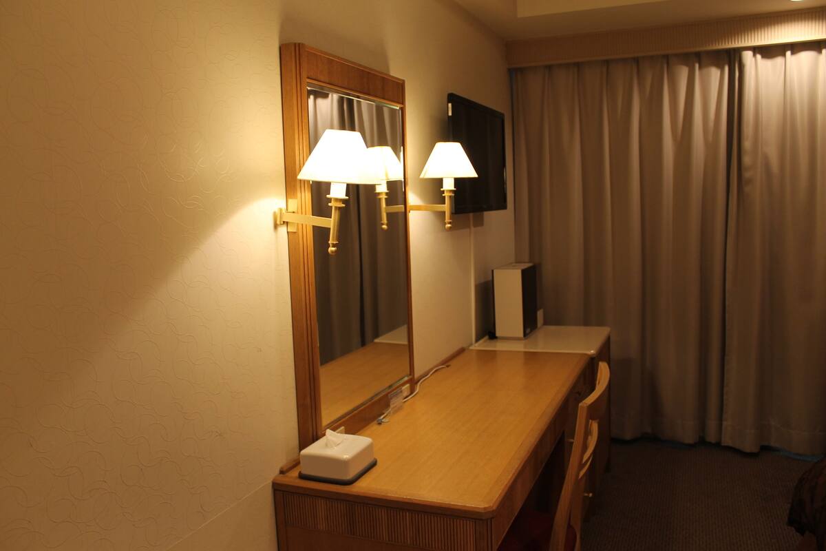 Une salle de bain d’hôtel avec un meuble-lavabo en bois, deux lampes fixées au mur, un miroir, un téléviseur et une fenêtre avec des rideaux.