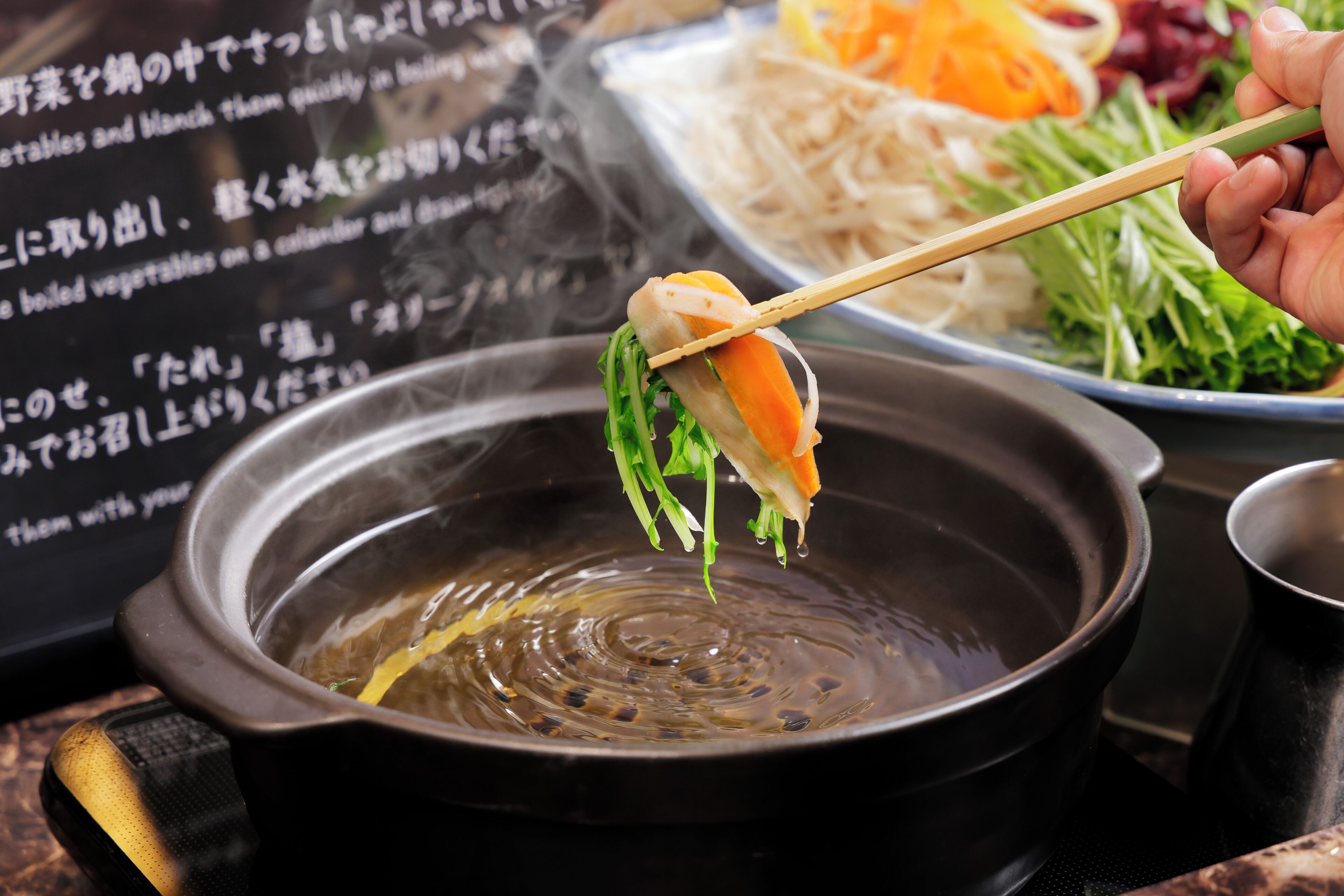 A bowl of soup with chopsticks holding a piece of food, a plate of noodles, and a menu in the background.