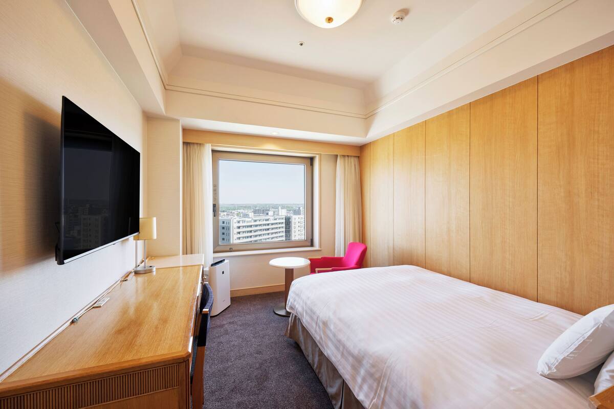Une chambre d’hôtel comprenant un lit, un bureau avec un téléviseur à écran plat, une petite table ronde, une chaise et une fenêtre donnant sur des immeubles.