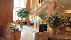 Un vestíbulo de hotel con una mesa de recepción, una planta grande en maceta, una pantalla digital y una escalera que sube a un nivel superior.
