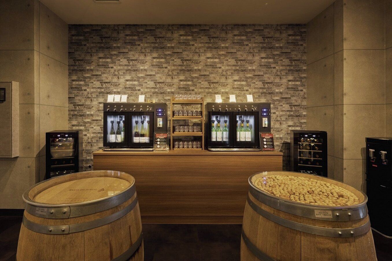 Una zona de bar con dos barriles de madera, una pared de ladrillo y dos refrigeradores con botellas.