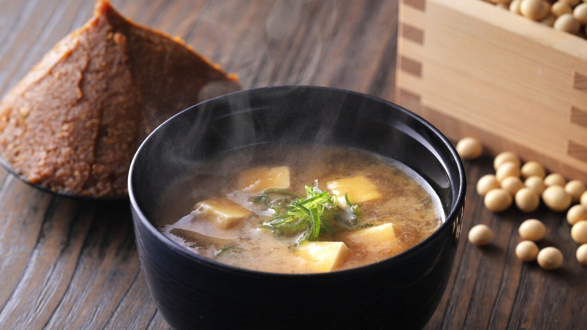 A bowl of soup with tofu and green vegetables, accompanied by a piece of bread and soybeans on a wooden surface.