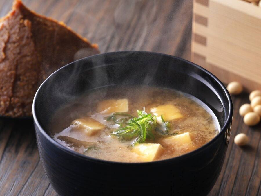A bowl of soup with tofu and green vegetables, accompanied by a piece of bread and soybeans on a wooden surface.