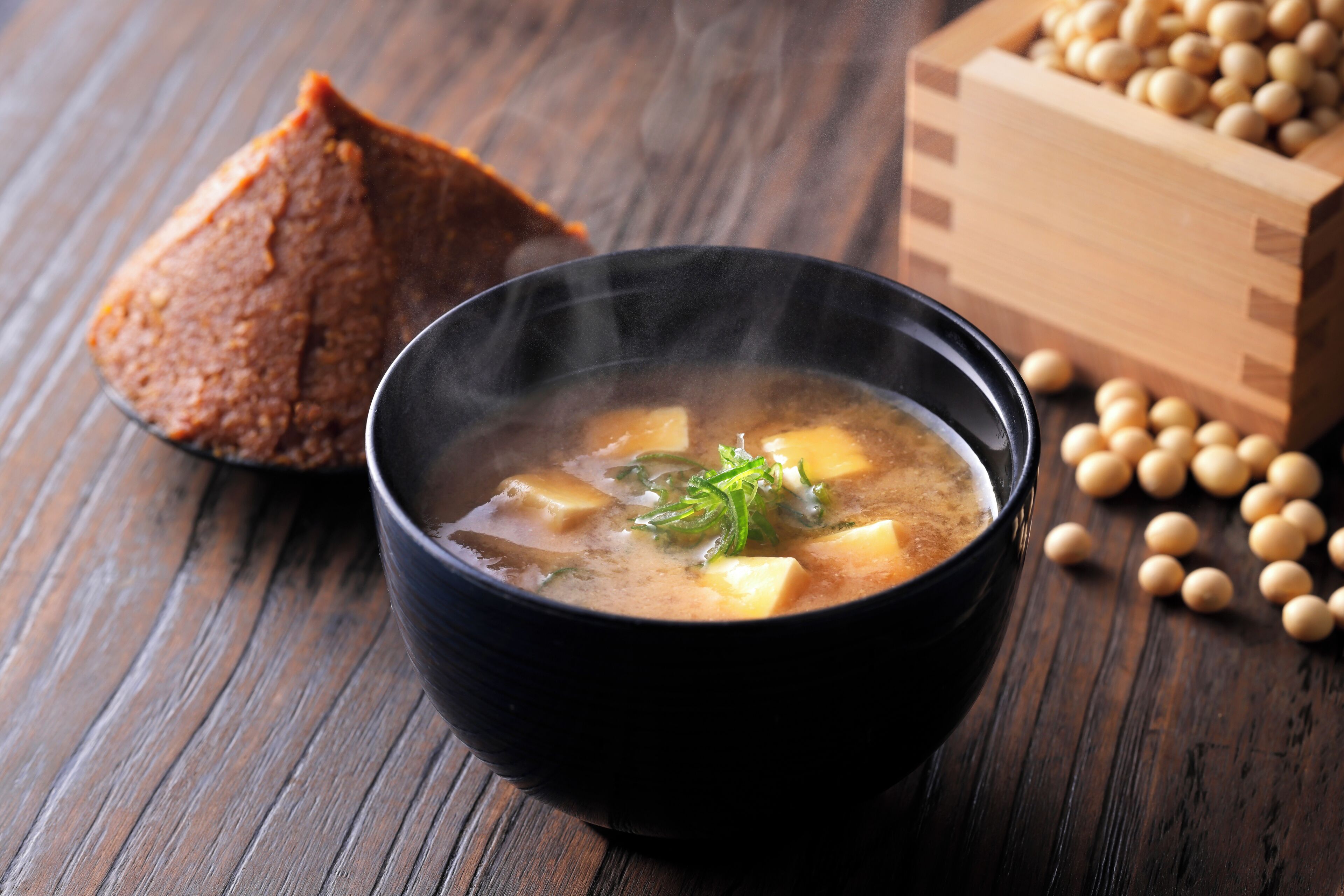 A bowl of soup with tofu and green onions, accompanied by a piece of bread and a wooden box of soybeans on a wooden table.