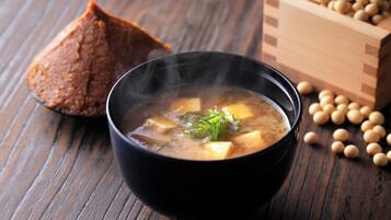 A bowl of soup with tofu and green onions, accompanied by a piece of bread and a wooden box of soybeans on a wooden table.