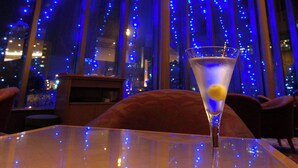 Una copa de martini sobre una mesa, con vista a la ciudad a través de amplios ventanales decorados con luces azules.