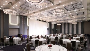 Une vaste salle de banquet, avec des tables rondes dressées pour le repas, agrémentée de lustres et dotée d’une scène.