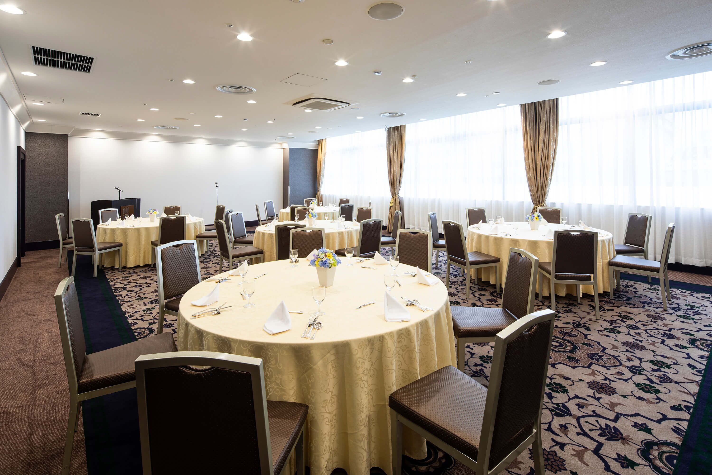 Un salón de conferencias con varias mesas redondas, cada una con sillas, manteles blancos y centros de mesa florales.